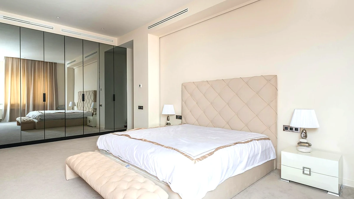 Tufted bench at the foot of a bed in a bright bedroom with bedside lamps and a white nightstand